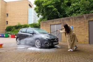 Person washing a black car with a power hose outside a building